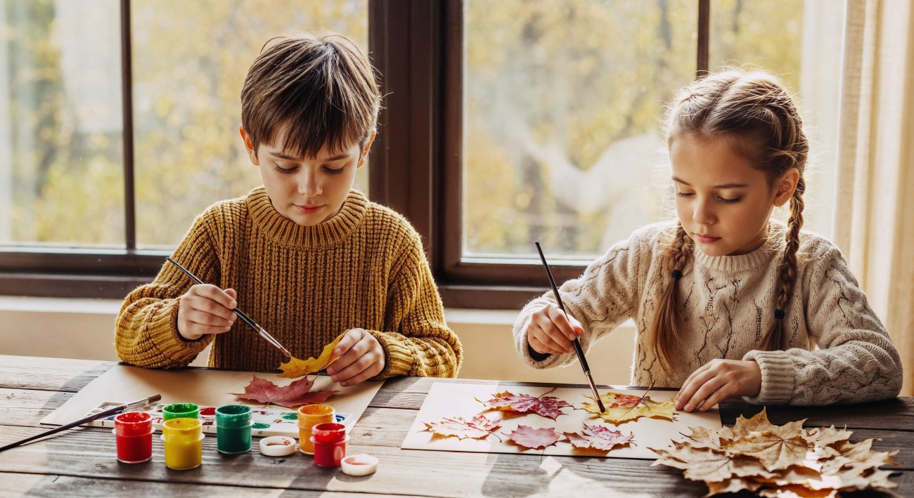 Deux enfants peignant des feuilles d’automne sur une table, avec des pots de peinture colorée devant une grande fenêtre lumineuse.