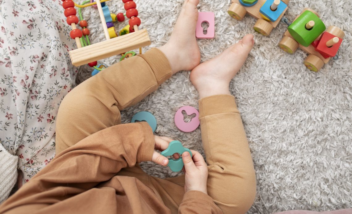 Pieds d’un bébé assis sur un tapis entouré de jouets en bois et d’une petite voiture
